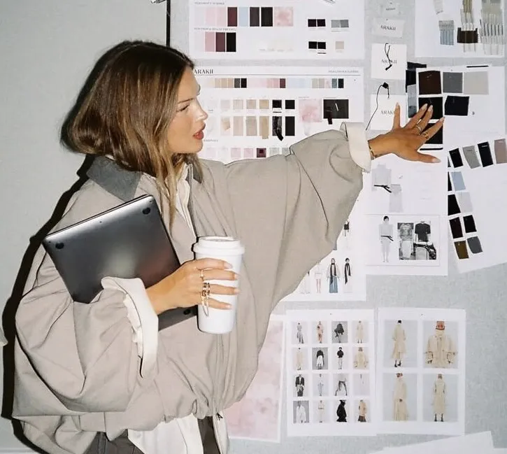 A designer holding a laptop and a coffee, reaching up to a pinned moodboard